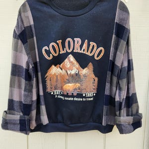 May include: A navy blue sweatshirt with the word "COLORADO" in orange lettering. The graphic features a mountain scene with a bear and trees. The sleeves are a grey and navy blue plaid pattern. The text "EST 1893" and "A strong, innate desire to travel" are also visible.