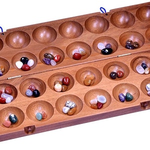 May include: A wooden Mancala board game with a hinged lid. The board is divided into two rows of 14 cups, each containing colorful stones. The board is made of light brown wood and has a natural finish.
