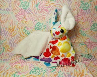 Peut inclure: Une chauve-souris en peluche blanche avec des cœurs arc-en-ciel et une aile en fourrure blanche. La chauve-souris est assise sur un tissu à motifs colorés.