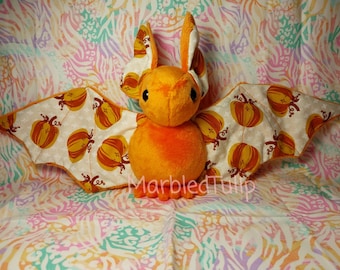 Peut inclure: Une peluche chauve-souris orange avec de grandes ailes ornées de citrouilles. La chauve-souris a de grandes oreilles et de petits yeux noirs. Les ailes sont de couleur crème avec des citrouilles orange et marron. Le texte "Marbled Tulip" est visible en bas.