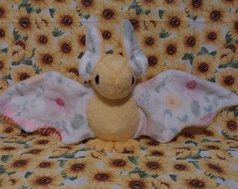 Peut inclure: Une peluche en forme de chauve-souris jaune avec des ailes à motifs floraux blancs. La chauve-souris a de grandes oreilles et de petits yeux noirs. Le fond est un motif de tournesols.