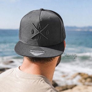 Könnte beinhalten: Eine anthrazitfarbene Snapback-Cap mit einem gestickten Design aus zwei gekreuzten Linien, den Buchstaben D, A, D und der Zahl 17. Ein silber-schwarzer Aufkleber auf dem flachen Schirm zeigt das Wort Snapback.