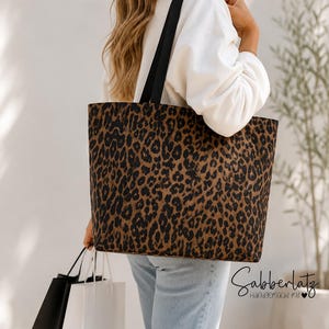 Könnte beinhalten: Eine große Tragetasche mit braun-schwarzem Leopardenmuster und zwei schwarzen Schulterriemen. Die Tasche ist rechteckig geformt und trägt den Schriftzug Sabberlatz Handgemacht mit in schwarzer Schreibschrift auf der unteren rechten Seite.