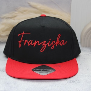 May include: Black and red snapback hat with the name "Franziska" embroidered in red script. The hat has a flat brim and a red button on top. The hat is a stylish accessory.