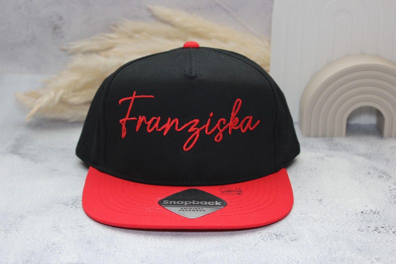 May include: Black and red snapback hat with the name "Franziska" embroidered in red script. The hat has a flat brim and a red button on top. The hat is a stylish accessory.