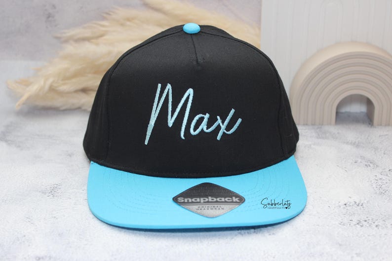 May include: Black and light blue Snapback hat with the name "Max" embroidered in light blue script. The hat features a flat brim and a snap closure. A small, light blue button is on top of the hat.