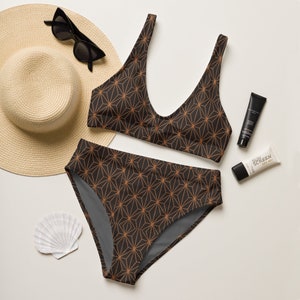 Puede incluir: Un conjunto de bikini de dos piezas con un estampado geométrico en negro y cobre. La imagen también incluye un sombrero para el sol, gafas de sol, protector solar y una concha. El top tiene cuello redondo y la braguita es de cintura alta.