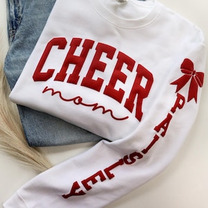 May include: White sweatshirt with the words "CHEER mom" in red, with a red bow and the word "PAISLEY" on the sleeve. The sweatshirt is folded on top of a pair of blue jeans.