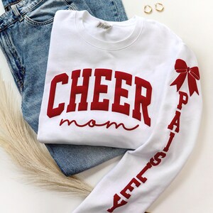 May include: White sweatshirt with the words "CHEER mom" in red block letters across the chest. The right sleeve features the name "PAISLEY" in red, with a red bow graphic. The sweatshirt is paired with blue jeans and gold hoop earrings.