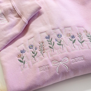 May include: A light pink sweatshirt with the word "MAMA" embroidered in white block letters, each letter adorned with floral sprigs. The sleeve has the name "Matilda" embroidered. A bow and the year "2025" are also embroidered.