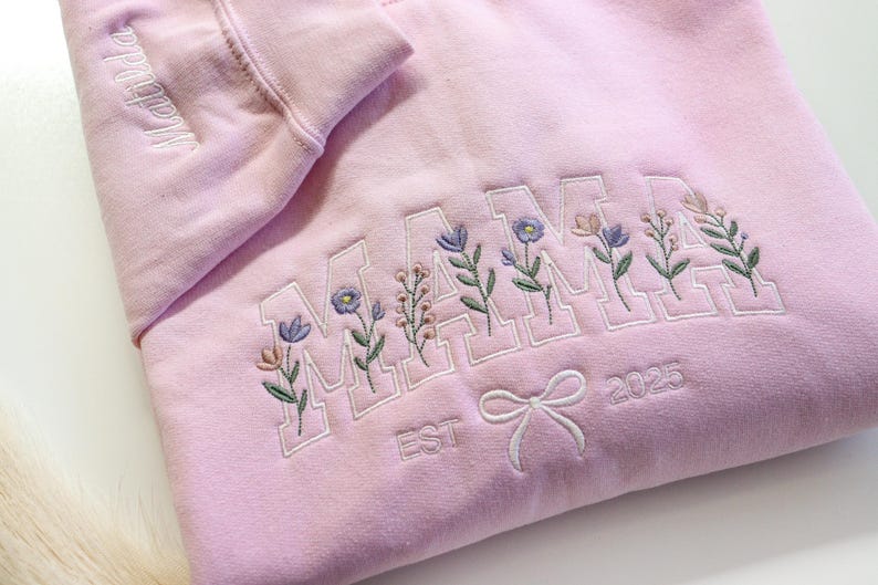 May include: A light pink sweatshirt with the word "MAMA" embroidered in white block letters, each letter adorned with floral sprigs. The sleeve has the name "Matilda" embroidered. A bow and the year "2025" are also embroidered.