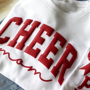May include: White sweatshirt with the word "CHEER" in large, red, block letters and the word "mom" in a red script font. A red bow is visible on the sleeve. The sweatshirt is likely for a cheerleading mom.