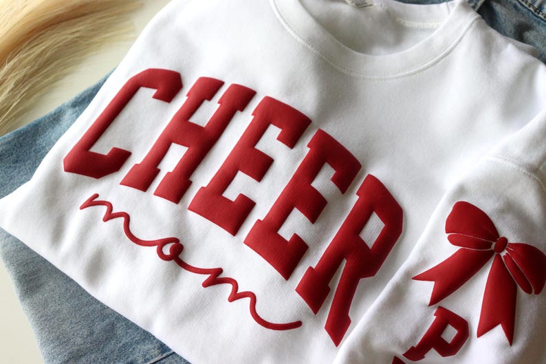 May include: White sweatshirt with the word "CHEER" in large, red, block letters and the word "mom" in a red script font. A red bow is visible on the sleeve. The sweatshirt is likely for a cheerleading mom.