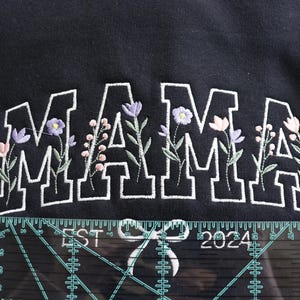 May include: Black sweatshirt with the word "MAMA" embroidered in white block letters, accented with floral designs in purple, pink, and green. The text "EST 2024" is also embroidered below the word "MAMA".