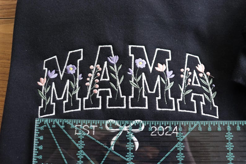 May include: Black sweatshirt with the word "MAMA" embroidered in white block letters, accented with floral designs in purple, pink, and green. The text "EST 2024" is also embroidered below the word "MAMA".