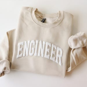 May include: A beige sweatshirt with the word "ENGINEER" in white, arched lettering across the front. The sleeves are cuffed, and the garment is folded. The sweatshirt is made of a soft material.