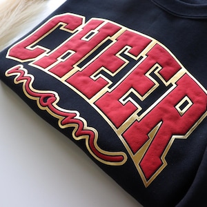 May include: A navy blue sweatshirt featuring the word "CHEER" in large red letters with gold edging. The word "mom" is written in gold script below. The sweatshirt is folded.