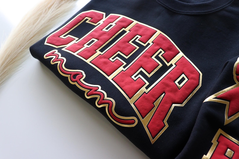 May include: A navy blue sweatshirt featuring the word "CHEER" in large red letters with gold edging. The word "mom" is written in gold script below. The sweatshirt is folded.