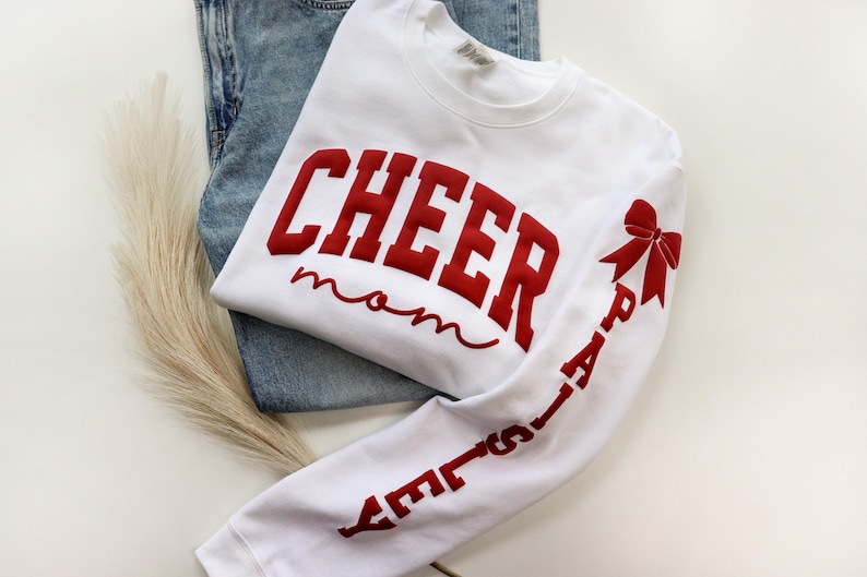 May include: White sweatshirt with the words "CHEER mom" in red, with a red bow and the word "PAISLEY" on the sleeve. The sweatshirt is folded on top of a pair of blue jeans.