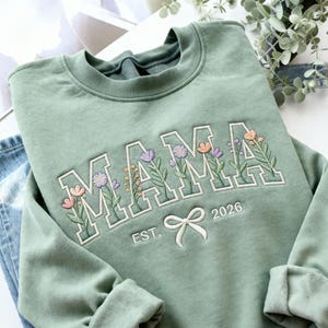 Könnte beinhalten: Salbeigrünes Sweatshirt mit dem gestickten Wort "MAMA" in weißen Blockbuchstaben, jeder Buchstabe mit bunten Blumenmustern gefüllt. Darunter eine weiße Schleife und der Text "EST. 2026".