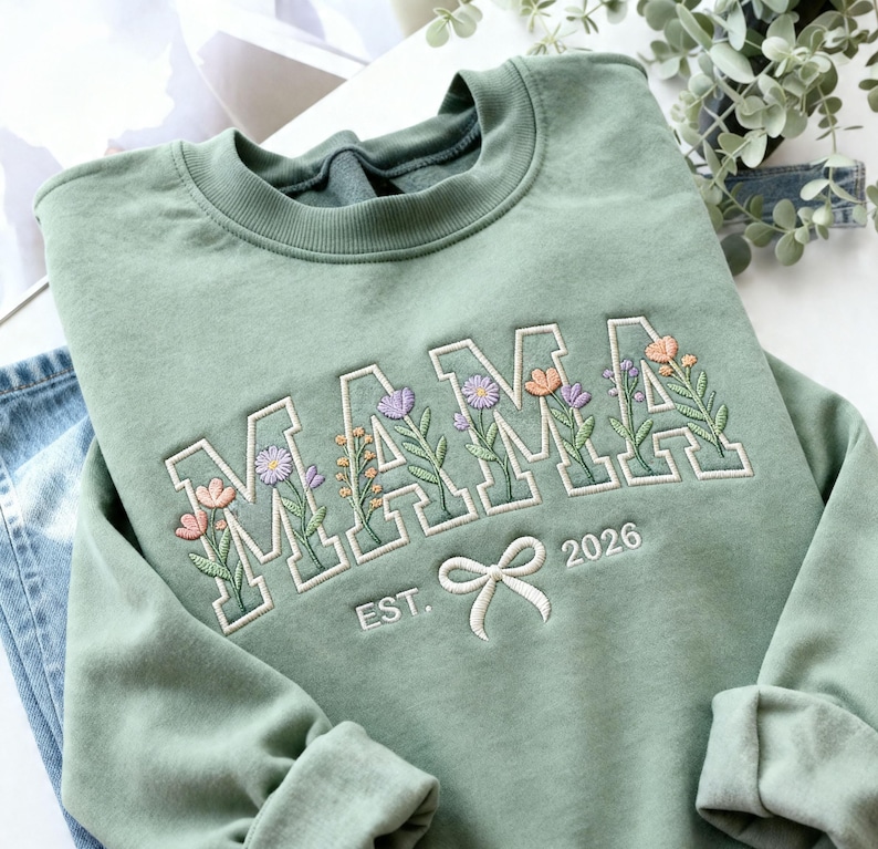 May include: Sage green sweatshirt with the embroidered word "MAMA" in white block letters, each letter filled with colorful floral designs. Below the word is a white bow and the text "EST. 2026".