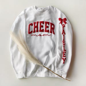 May include: White sweatshirt with red embroidered text that reads "CHEER mom" across the chest. The right sleeve has the name "Paisley" in red lettering and a red bow. A dried, feathery plant stem lies across the sweatshirt.