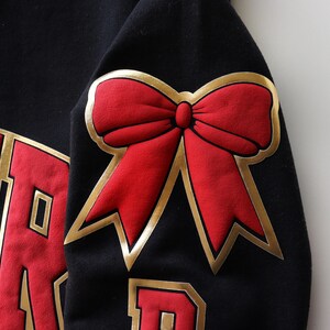 May include: Black sweatshirt with a large red bow with gold trim on the sleeve. The sweatshirt also features red lettering with gold trim. The bow is a prominent design element.