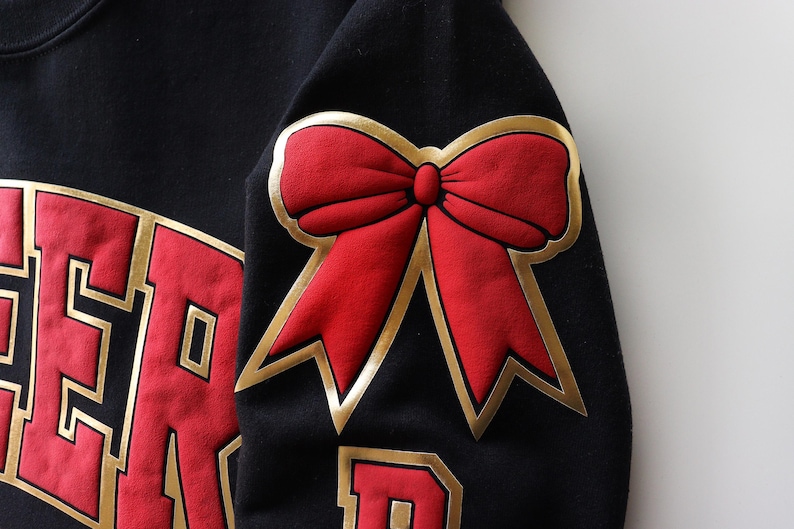 May include: Black sweatshirt with a large red bow with gold trim on the sleeve. The sweatshirt also features red lettering with gold trim. The bow is a prominent design element.