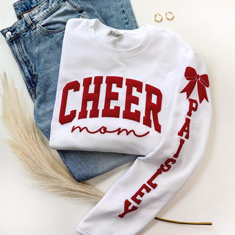 May include: White sweatshirt with the words "CHEER mom" in red block letters across the chest. The right sleeve features the name "PAISLEY" in red, with a red bow graphic. The sweatshirt is paired with blue jeans and gold hoop earrings.