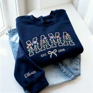 May include: A navy blue sweatshirt with the word "MAMA" embroidered in white block letters, adorned with floral accents. Below the word "MAMA" is the text "EST. 2026" and a bow. The sleeve has the name "Olivia" embroidered. The sweatshirt is paired with blue jeans.