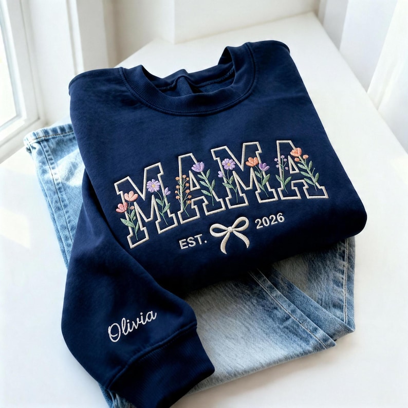 May include: A navy blue sweatshirt with the word "MAMA" embroidered in white block letters, adorned with floral accents. Below the word "MAMA" is the text "EST. 2026" and a bow. The sleeve has the name "Olivia" embroidered. The sweatshirt is paired with blue jeans.