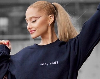 May include: A woman wearing a dark blue oversized sweatshirt with the text "yes, and?" embroidered on the front. She is also wearing blue jeans and a brown leather handbag.