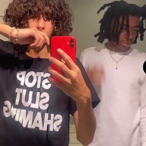 May include: Two people, one wearing a black t-shirt with the text "SHAMING SLUT STOP" in white, and the other wearing a white long-sleeved shirt, are standing in a bathroom. The person in the black t-shirt is holding a red iPhone.