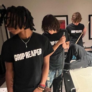 May include: Several people wearing black t-shirts with the text "GRIP REAPER" in white, gathered around a pool table. One person is holding a pool cue. The scene is indoors, with a neutral-toned background.