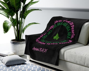 Può includere: Una coperta nera drappeggiata su un divano bianco. La coperta presenta un design circolare con il testo "Pretty Girls Wear 20 Pearls. AKA AKA AKA" in rosa e verde. Un trifoglio verde è al centro e il testo "Amber FA 21'" è sotto.