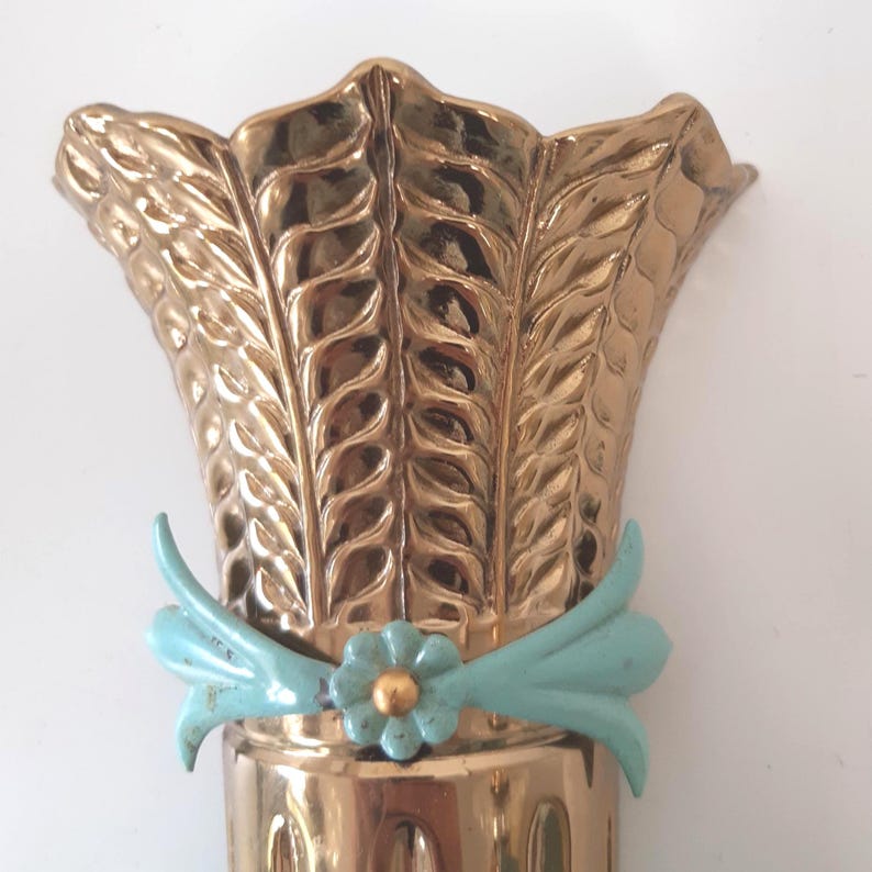 May include: A gold-colored metal decorative vase with a wheat sheaf design. The vase has a light blue bow and a flower with a gold center. The vase is likely for decorative purposes.