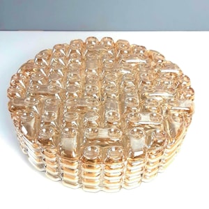 May include: A round, amber-colored glass light fixture with a textured, woven pattern. The fixture has a series of raised, rectangular blocks arranged in a grid-like design. The glass has a slight iridescence, reflecting light.