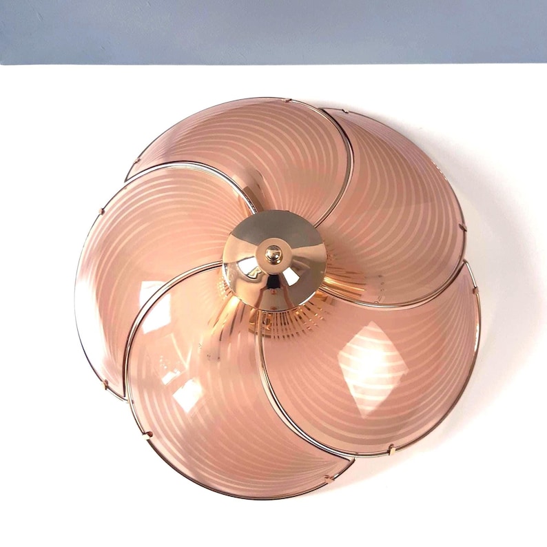 May include: A decorative ceiling light fixture with a flower-like design. The fixture features six petal-shaped, light pink glass shades with a striped pattern, held together by a gold-colored metal frame. The center is a gold-colored metal disc.