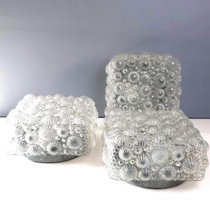 Puede incluir: Dos objetos decorativos de vidrio transparente con un patrón texturizado similar a flores. Uno tiene forma baja y rectangular, y el otro es más alto y vertical. Ambos se asientan sobre una base gris circular. El vidrio tiene una apariencia burbujeante y texturizada.