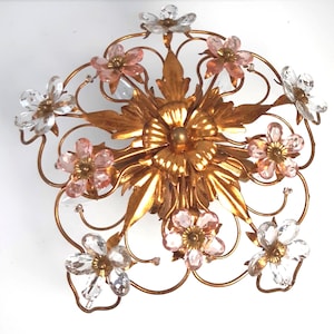 May include: A gold-toned metal wall sconce with a floral design. The sconce features a central flower with surrounding leaves and delicate, curved metalwork. It is decorated with pink and clear crystal flower accents, creating a vintage aesthetic.