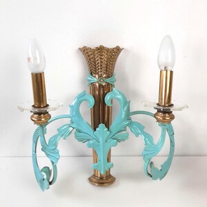 May include: A vintage wall sconce with two light fixtures. The sconce features a gold-colored central column and teal-colored, ornate arms. The sconce has a decorative, gold-colored top and clear glass candle holders. The light bulbs are white.