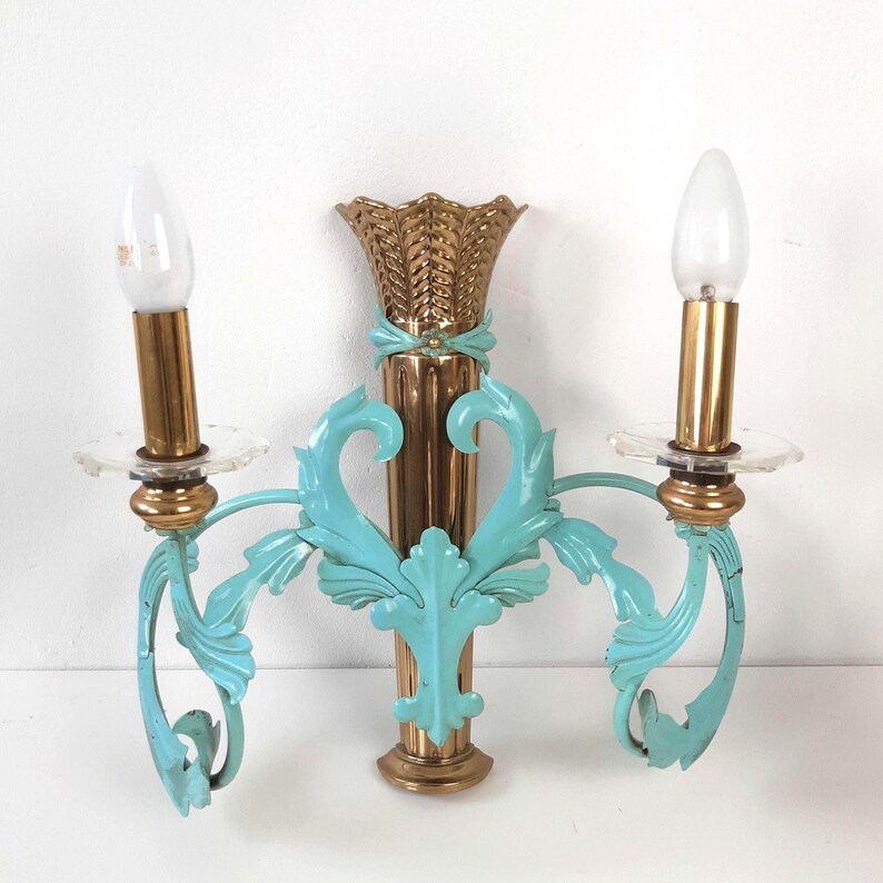 May include: A vintage wall sconce with two light fixtures. The sconce features a gold-colored central column and teal-colored, ornate arms. The sconce has a decorative, gold-colored top and clear glass candle holders. The light bulbs are white.