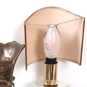 May include: A vintage wall sconce featuring a cream-colored lampshade with a gold trim. The sconce has a gold-colored base and a clear lightbulb. The design is simple and elegant, suitable for various interior styles.