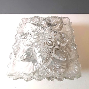 Può includere: Un apparecchio di illuminazione decorativo in vetro trasparente con un motivo floreale. L'apparecchio ha una forma quadrata ed è ricoperto da petali di vetro strutturati e sovrapposti. Un fiore è centrato sulla parte superiore. Il vetro è trasparente.