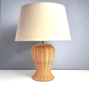 Könnte beinhalten: Eine Tischlampe aus Weide mit beigem Lampenschirm. Der Lampenfuß ist hellbraun und hat eine abgerundete Form. Der Lampenschirm ist hellbeige und hat eine klassische, konische Form. Die Lampe steht auf einer weißen Oberfläche.