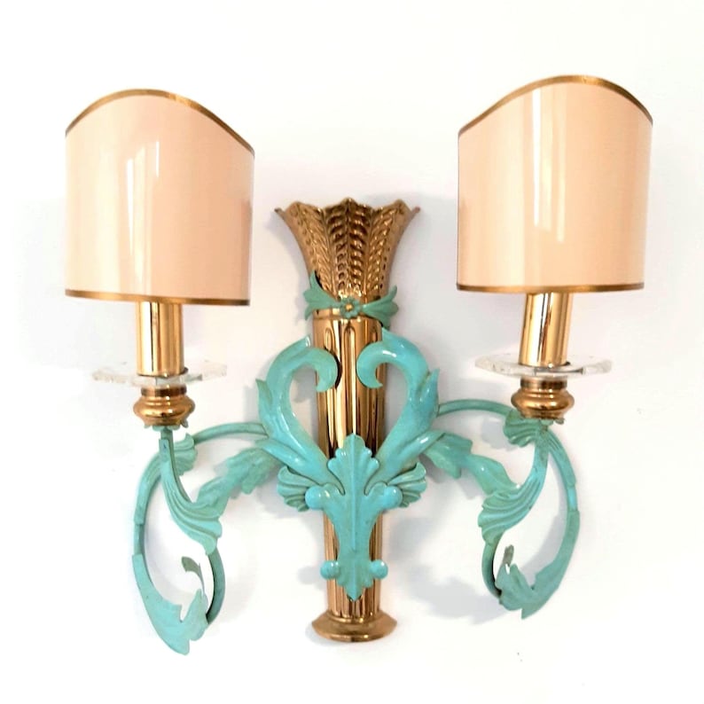 May include: A pair of wall sconces with a gold finish, teal accents, and cream-colored lampshades. The sconces feature a decorative design with a central column and two curved arms. The lampshades are a rounded shape with gold trim.