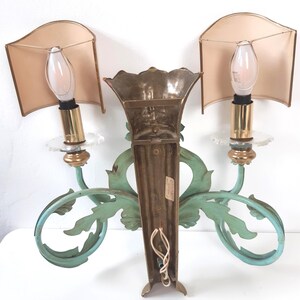 May include: Vintage wall sconce with two lights. The sconce features a central brass element and two light fixtures with beige shades. The sconce has a decorative, light-green, scroll-like design. The light fixtures have a clear, glass-like base.