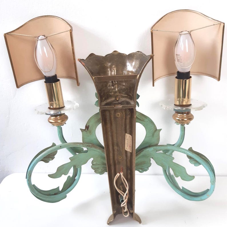 May include: Vintage wall sconce with two lights. The sconce features a central brass element and two light fixtures with beige shades. The sconce has a decorative, light-green, scroll-like design. The light fixtures have a clear, glass-like base.