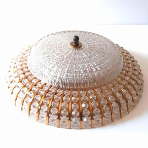 Op de afbeelding: Een vintage plafondlamp met een heldere, getextureerde glazen koepel en een lager gedeelte versierd met rijen heldere, facetgeslepen kristallen kralen. De lamp heeft een goudkleurig metalen frame en een kleine, donkerbruine houten finial.