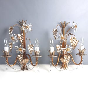 May include: Pair of gold-toned wall sconces with floral and leaf accents. Each sconce features clear crystal flower-shaped embellishments, leaf details, and dangling crystal strands. The sconces have two light bulb fixtures each.